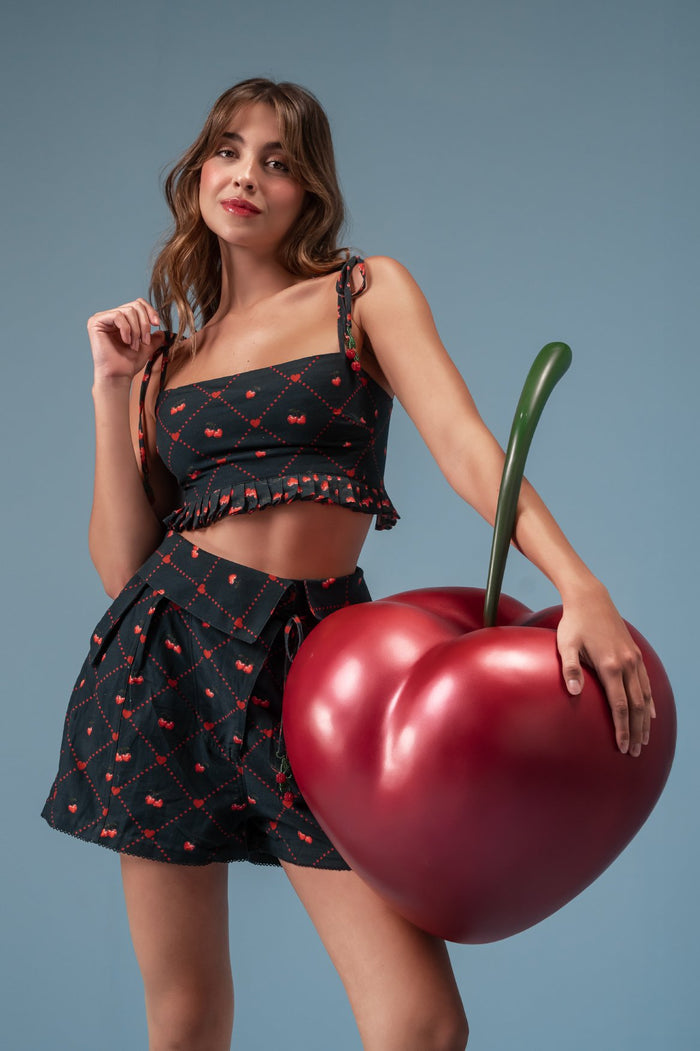 Black crop top with red cherry print in diagonal grid pattern, featuring ruffled hem and adjustable tie straps with cherry appliqués