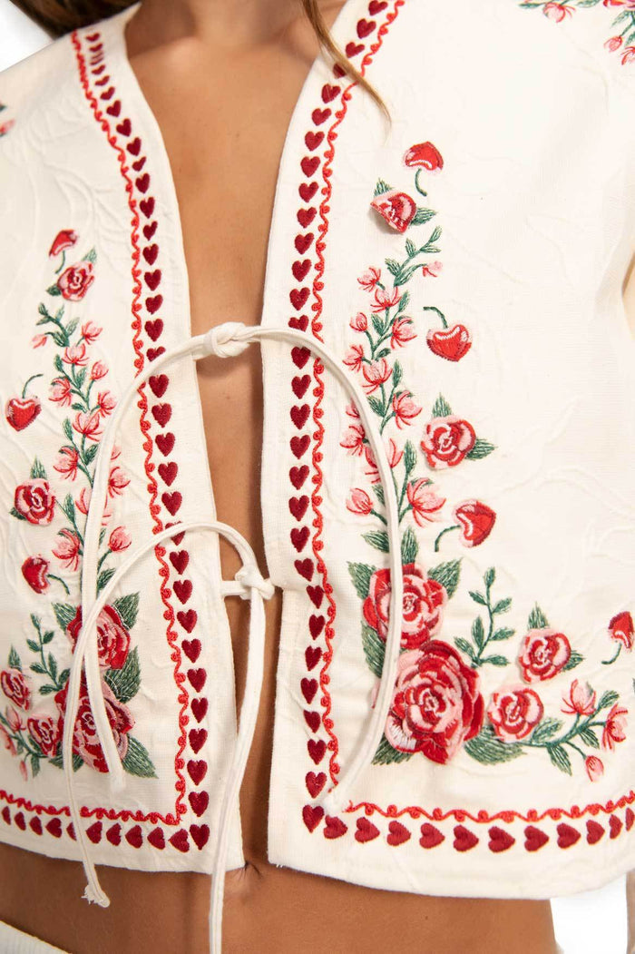 White cropped jacket with red heart embroidery trim and floral embroidery featuring roses and green foliage, tie-bow front closure