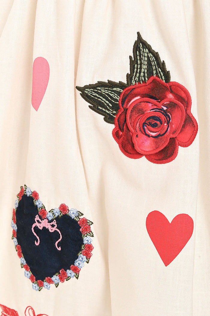 Cream girls' dress with embroidered red roses with black wings, pattern detail-framed hearts, and pink and coral heart appliqués