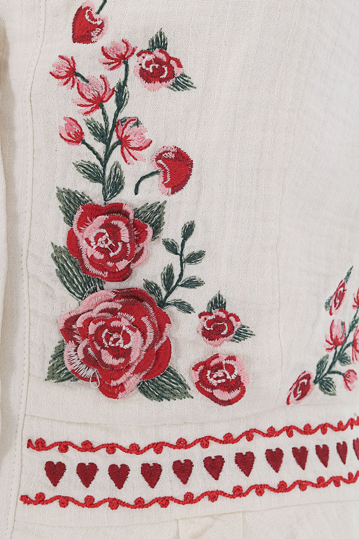 White shirt with red and pink embroidered floral detailing along the hem, featuring scattered blooms with green leaves and a decorative border of hea…