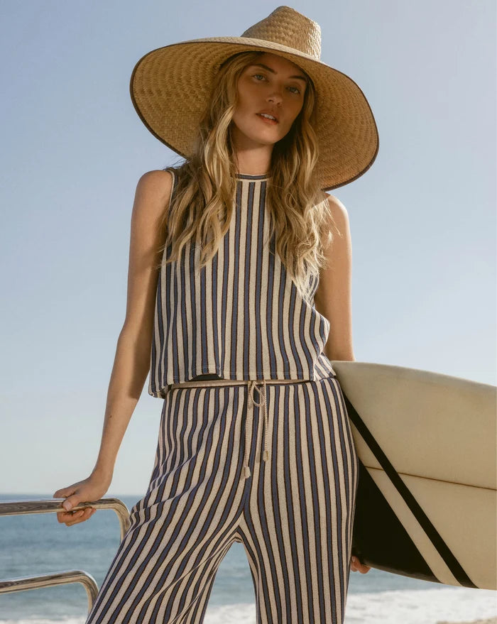 A woman in a striped outfit and wide-brimmed hat holds a surfboard against a backdrop of the ocean and clear sky.