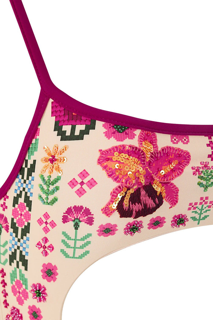 Magenta one-piece swimsuit with beige base, pixelated cross-stitch pattern of flowers, butterflies, and geometric shapes embellished with sequins and…
