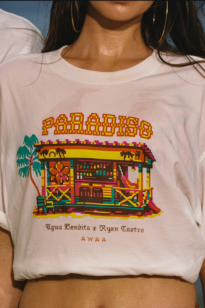 Cream t-shirt with pixelated paradise scene graphic featuring colorful beach hut, palm tree, and text reading Paradiso and Agua Bendita x Ryan Castro