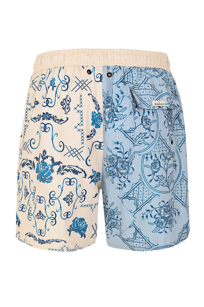 Men's swim trunks with split-panel design: cream side featuring blue scrollwork and cross-stitch embroidery pattern, light blue side with pattern det…
