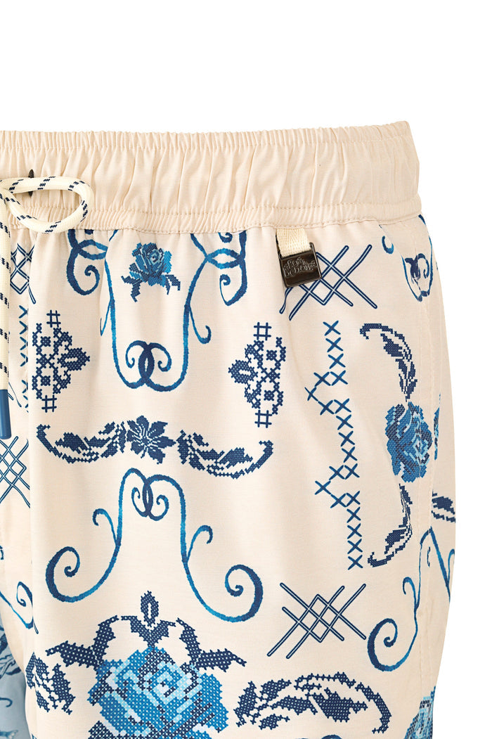 Cream men's trunks with blue embroidered-style print featuring scrollwork, cross-stitch patterns, and lattice details