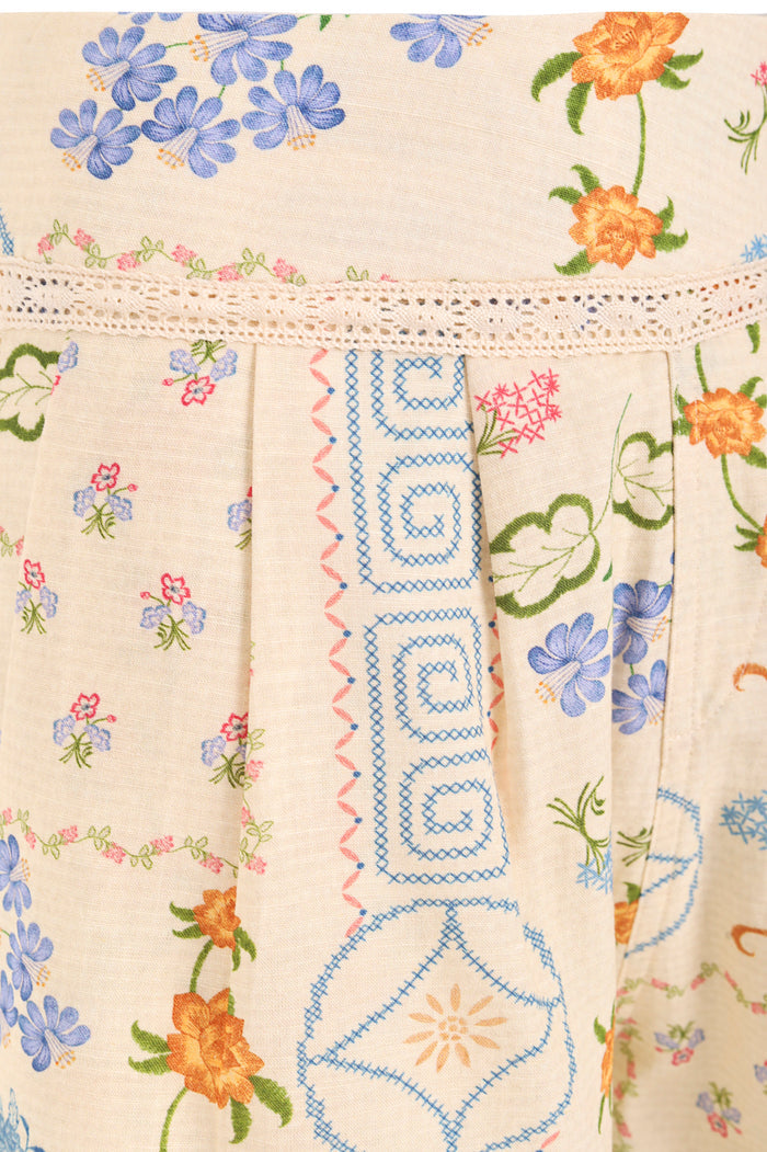 Cream shorts with embroidered pattern detail print in blue, orange, and pink, featuring crochet lace trim at waist and blue cross-stitch detailing on…