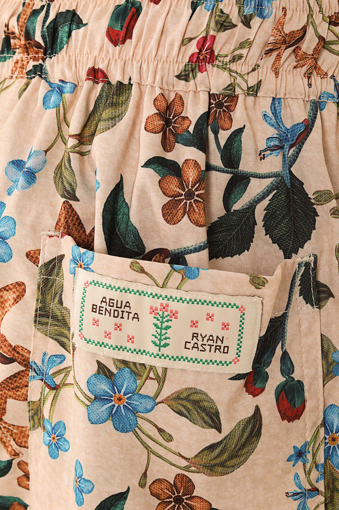 Beige swim trunks with botanical floral print in blue, orange, and green tones, elastic waistband, and embroidered label detail