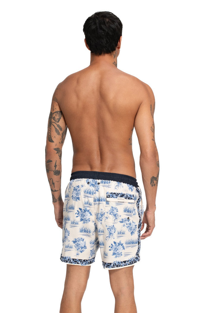 White swim trunks with blue toile-style print featuring tropical scenes, navy elastic waistband and contrast trim at hem, back view