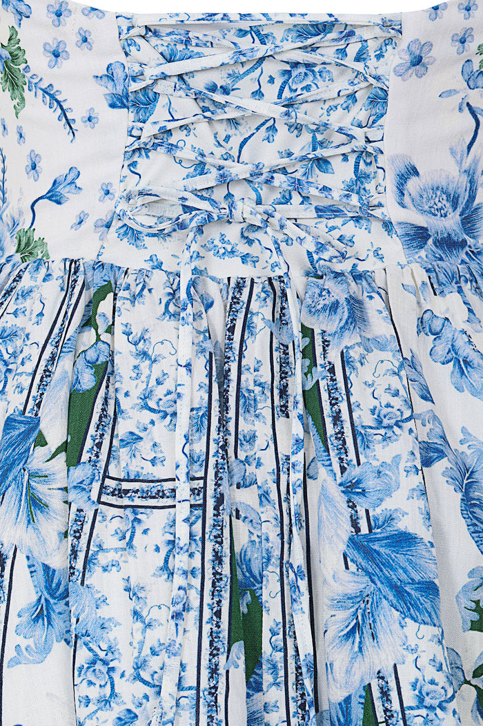 White dress with blue botanical print featuring ruched bodice with lace-up detail and pleated skirt with green accents