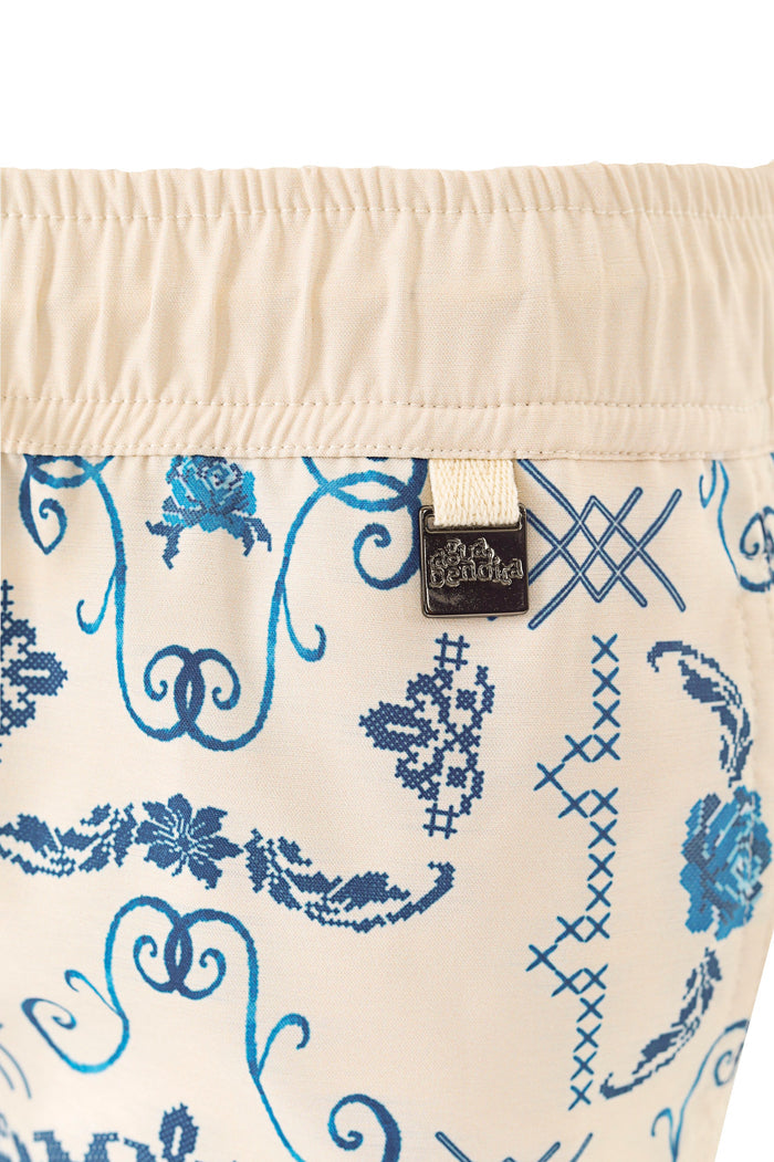 Cream swim trunks with blue embroidered print featuring scrolls, cross-stitch florals, and lattice details, elastic waistband with metal emblem