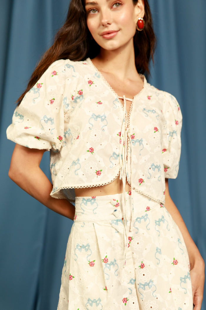 Cream eyelet crop shirt with blue scrollwork and pink pattern detail print, puffed sleeves, tie-bow front closure, and scalloped trim
