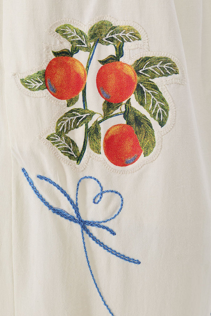 White linen pants with embroidered fruit and leaf design featuring red and orange round fruits with green foliage and blue decorative stitching